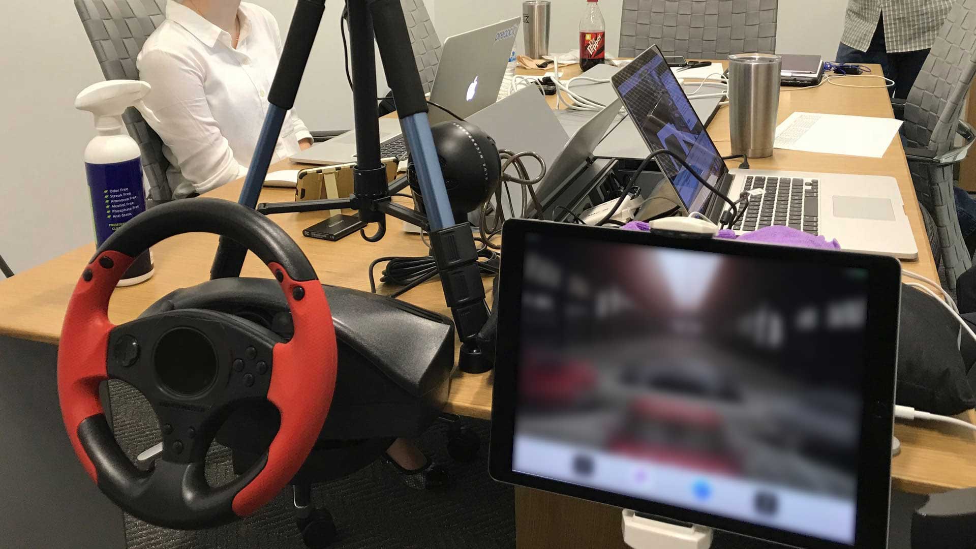 The live-streamed testing rig — a branded steering wheel and an iPad running the test prototype, wired to camera arms on stands and a bank of laptops for the moderator and remote observers.