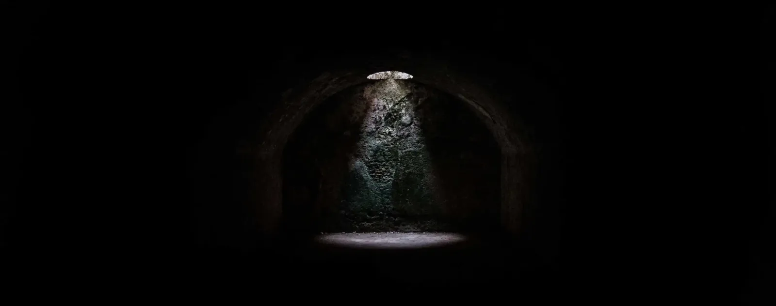 A dark vaulted stone chamber lit only by a narrow shaft of daylight falling from an opening in the ceiling onto the floor below.