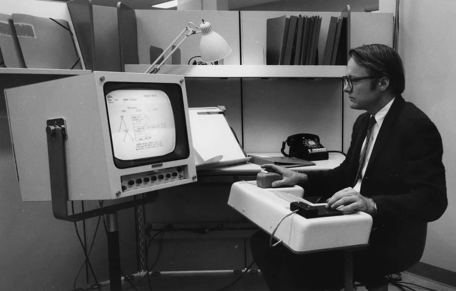 A researcher at a 1960s workstation with a CRT display, chord keyset, and the first computer mouse.