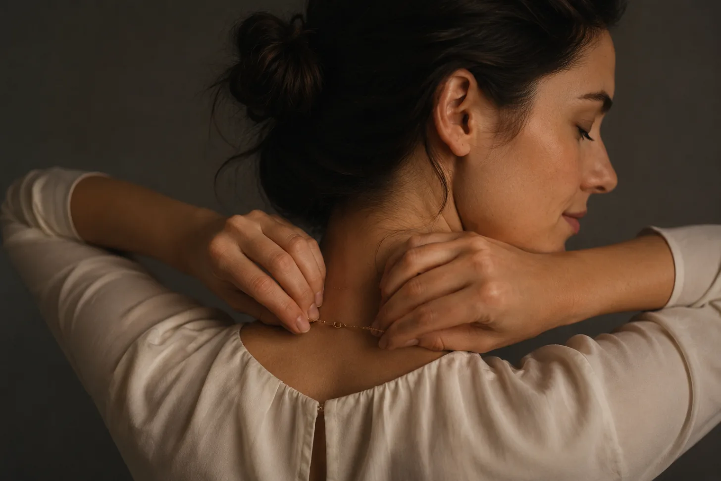 A woman reaching behind her neck to fasten a fine chain clasp she cannot see.
